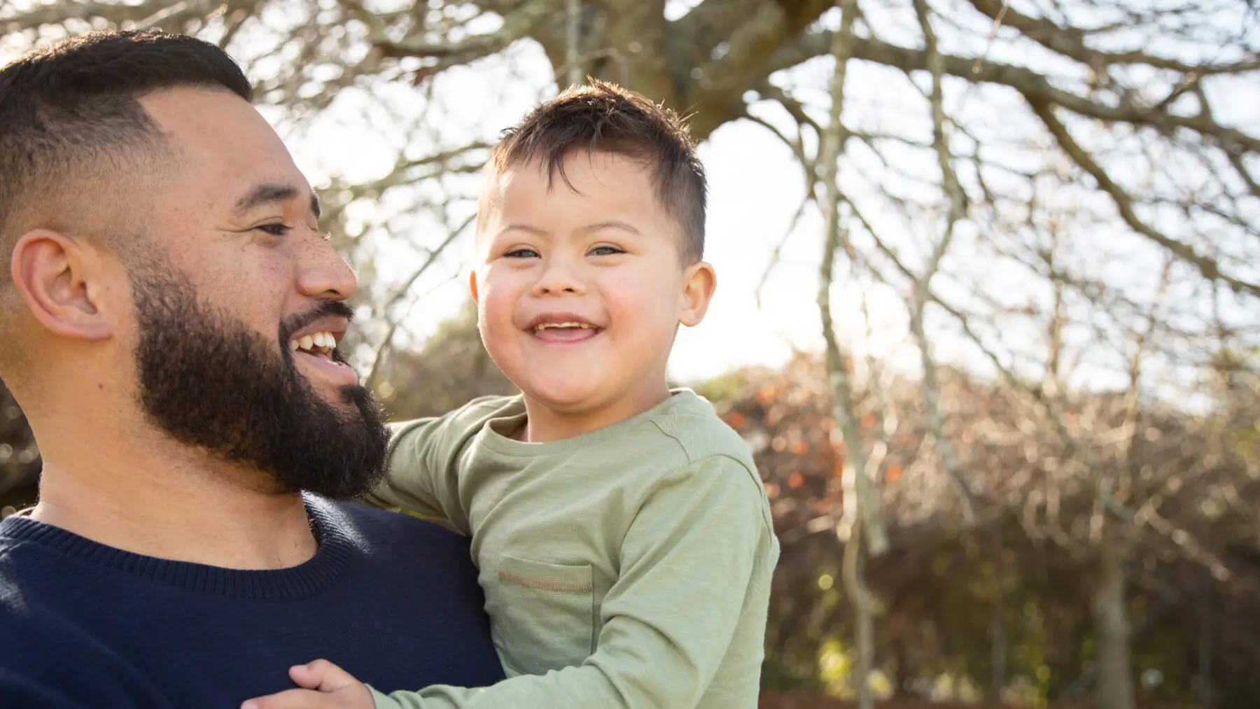 Father holds toddler son who has down syndrome outside in nature as both of them are smiling