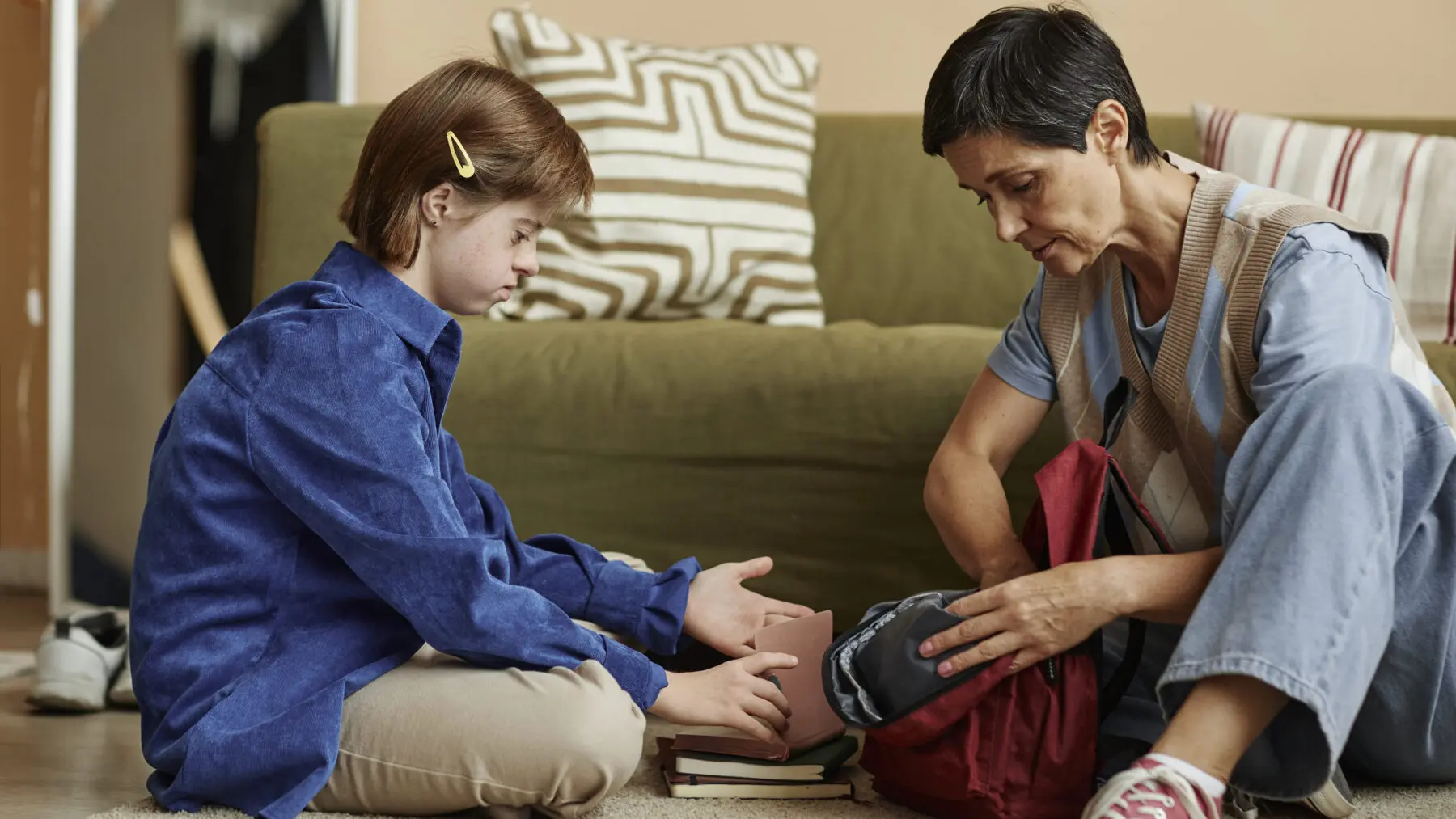 Woman and daughter with down syndrome packing backpack while seated on the floor.