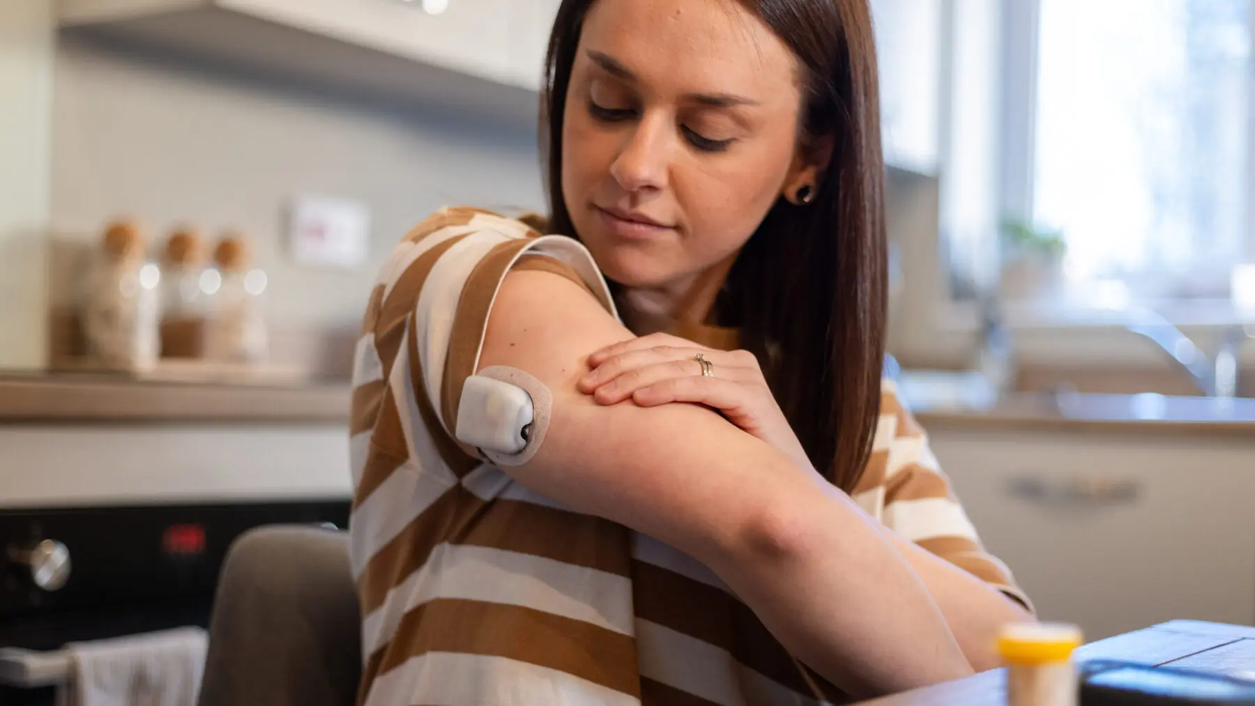Close-up of a woman sitting in her kitchen with her sleeve pulled up to reveal her Libre Continuous Glucose Monitor.