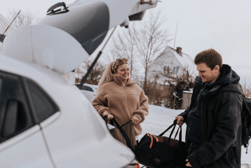 Woman smiling at man who is loading suitcase in car trunk with winter landscape behind them