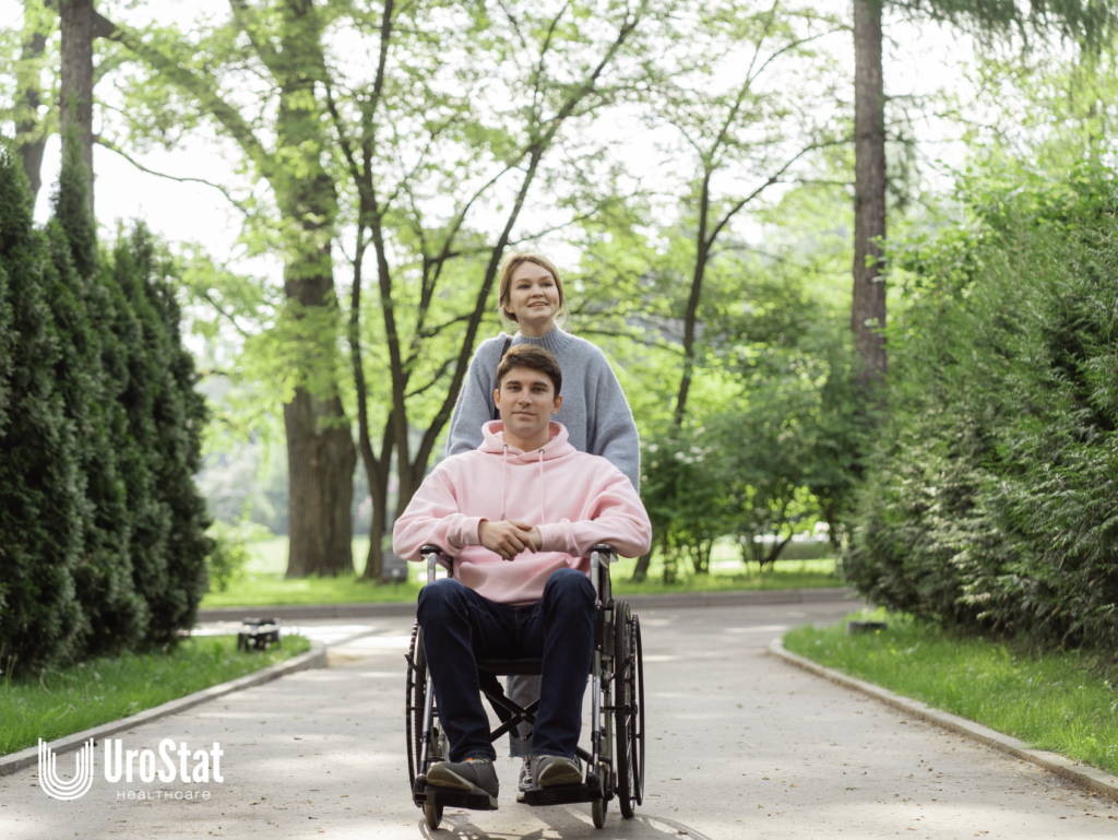A young man using a wheelchair on a park path with a woman standing behind him.