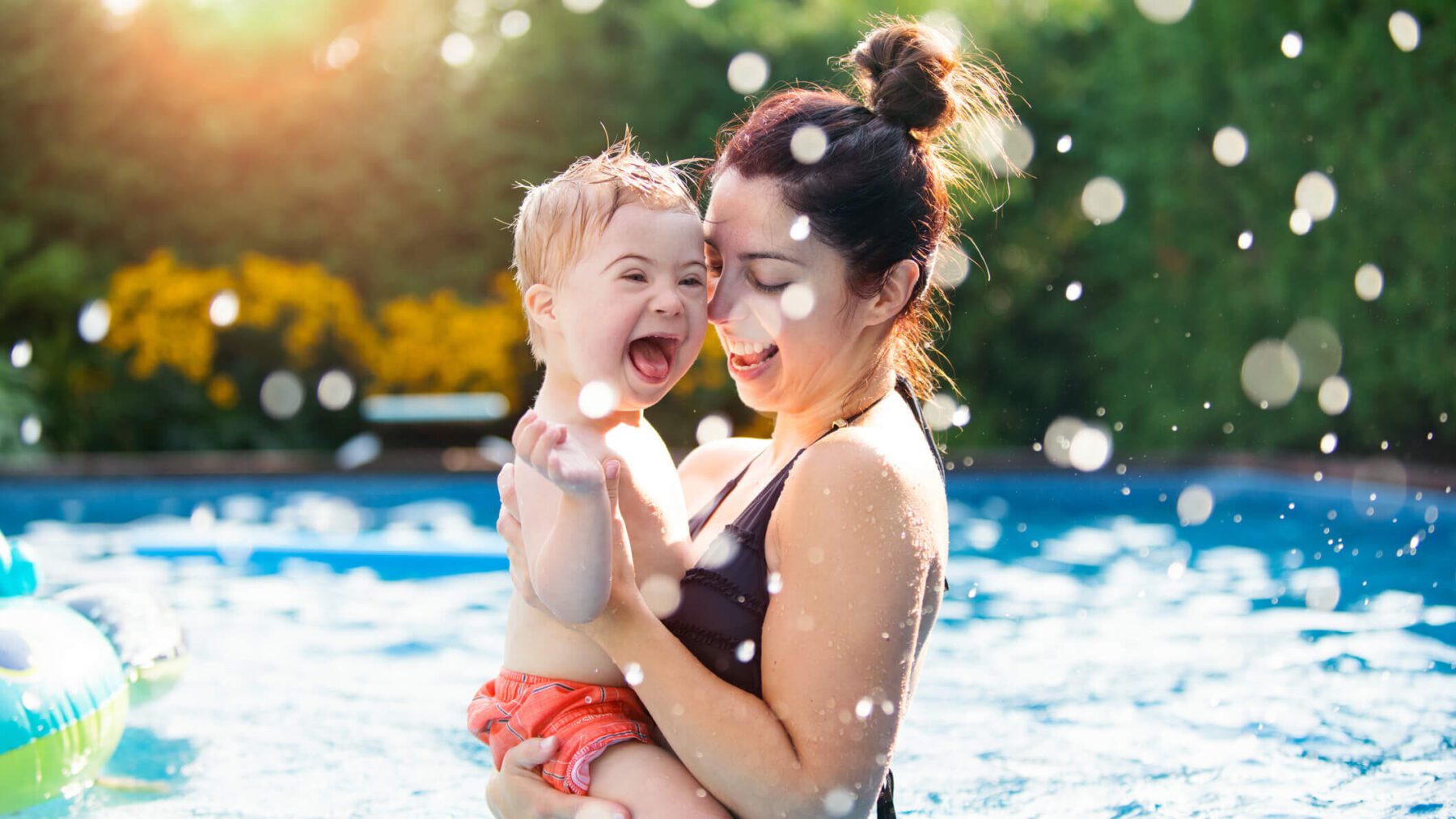 3-year-old little boy with down syndrome having fun in the swimming pool with his family with his mother holding him in her arms.