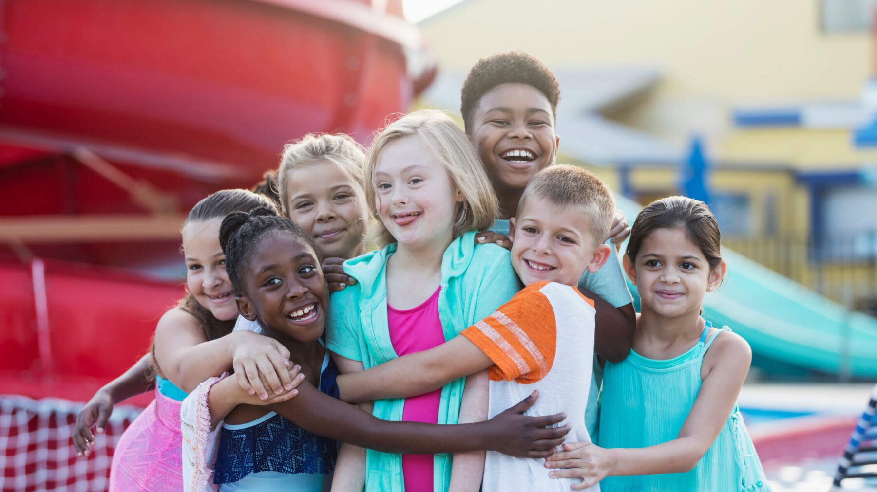 10 year old girl with down syndrome having fun at a water park, in the center of multi-ethnic group of friends, getting a group hug. All smiling at the camera.