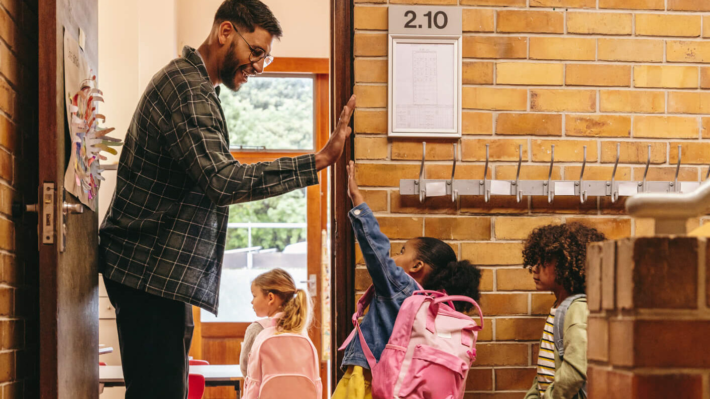 Male teacher greeting his elementary school students at the door to the classroom with a high five.