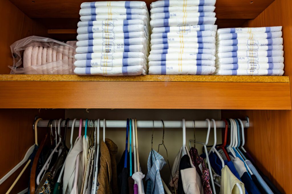 Clothing closet with clothes on hangers and a stack of adult diapers.
