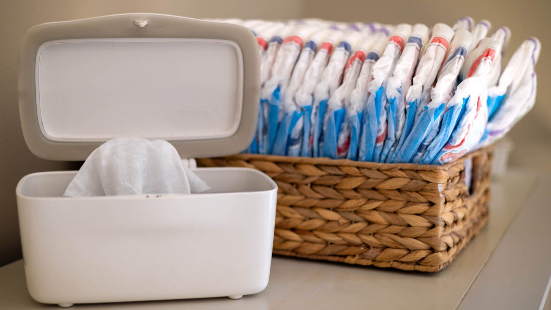 A diaper wipe bin open with a basket of diapers in the background.
