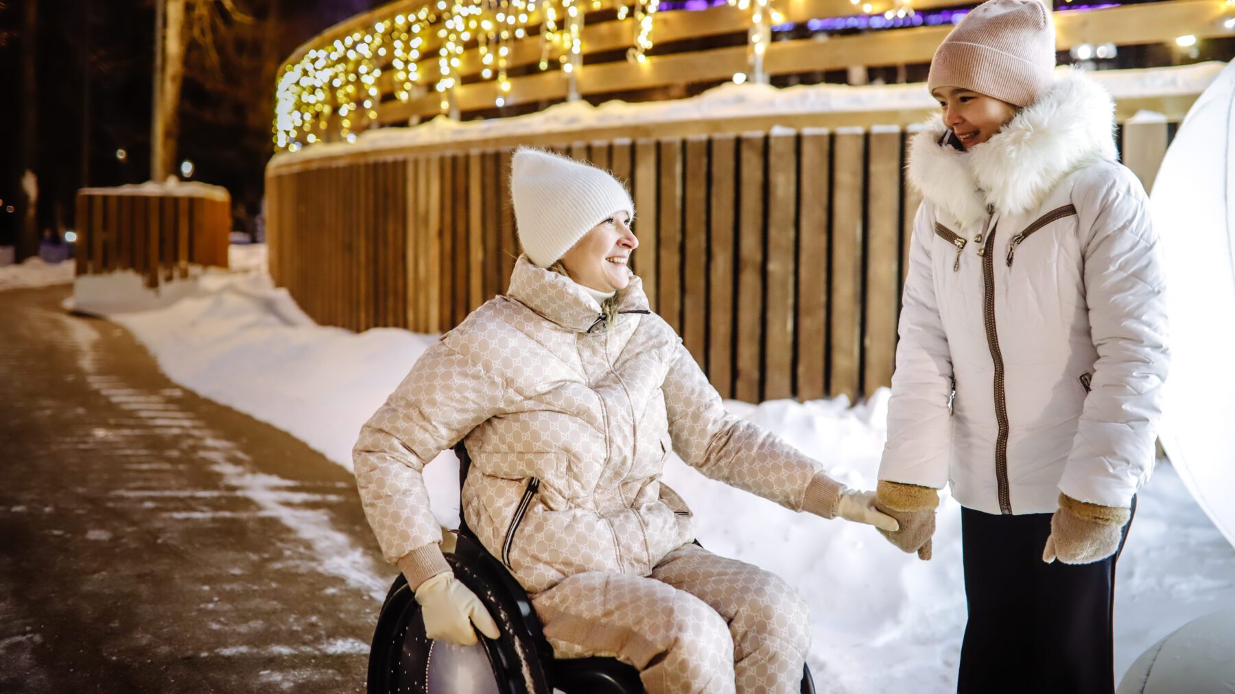 Mother in wheelchair with young daughter strolling in winter park with Christmas lights