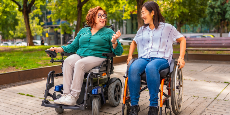 Two female friends in wheelchair outside in park smiling and talking