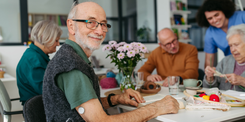 Elderly man smiling into camera at table with friends with CGM on the back of his arm