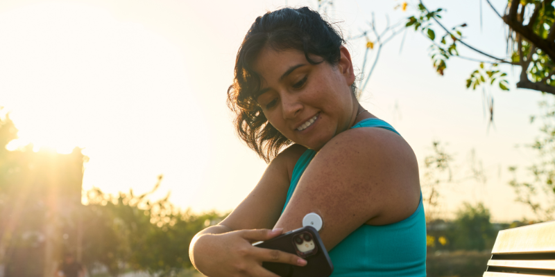 Woman sitting in workout clothes outdoor on a bench scanning her CGM with phone in the morning