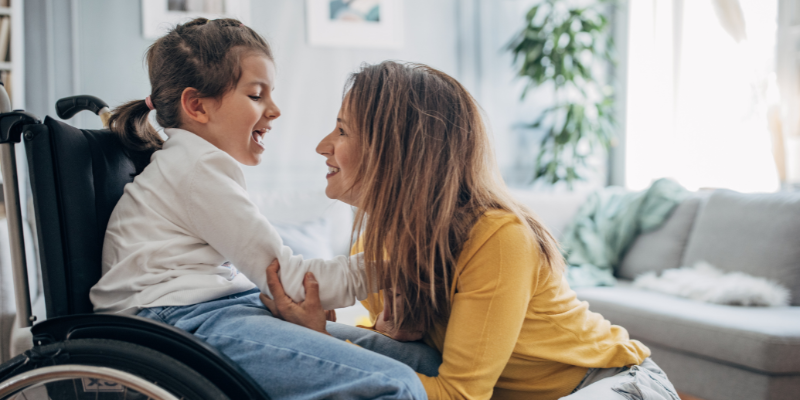 Daughter in wheelchair smiling at mom who is also smiling and kneeling in front of her