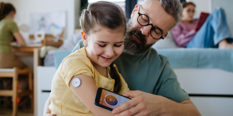 Father helping daughter check her blood sugar levels via CGM on her arm with smartphone