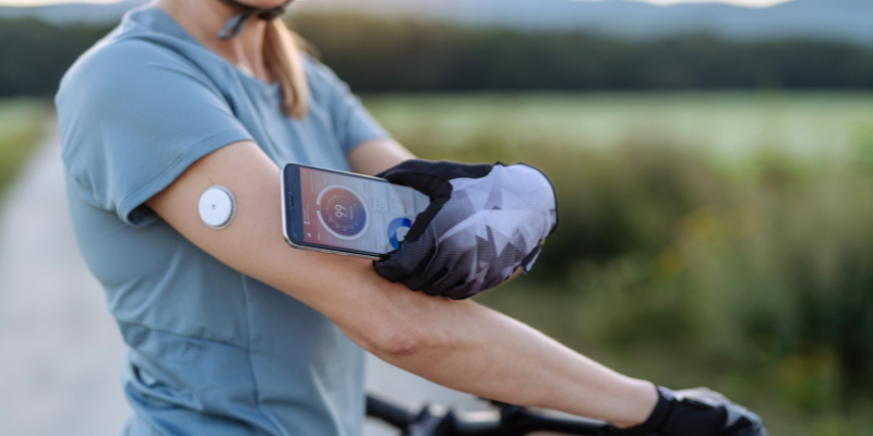 Woman biking with CGM on the back of her arm stops to check blood sugar with smartphone