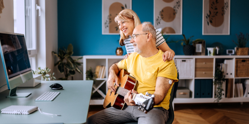 Older man with CGM on the back of his arm playing guitar in front of computer with his wife smiling next to him