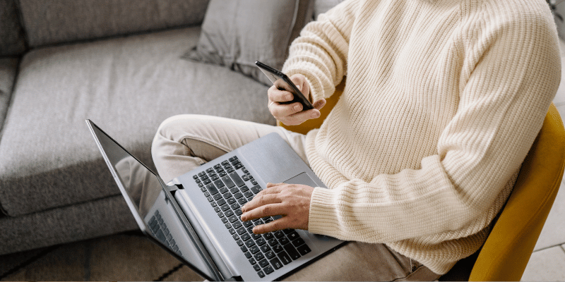 Image shows seated man typing on laptop and holding phone