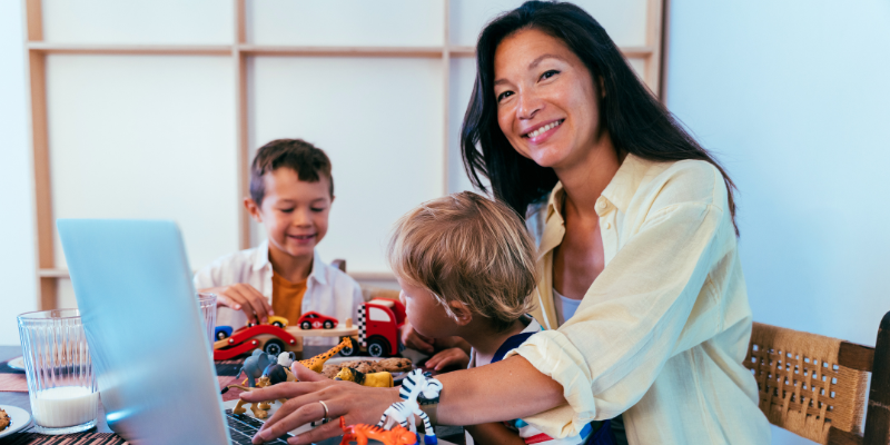 Mom smiling and using laptop with son on her lap and other son playing with cars next to them