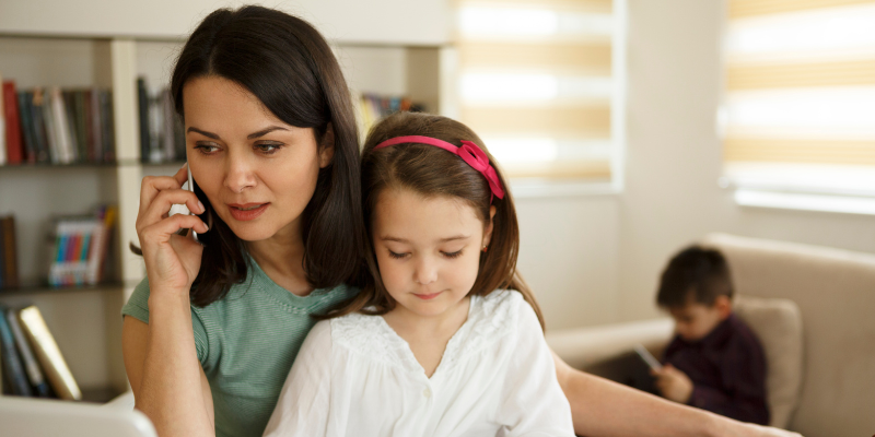 Mom on the phone with questions while daughter sits in her lap and son is in the background on tablet