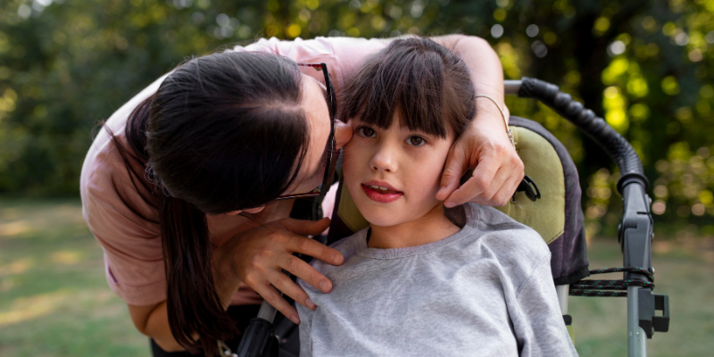 Mom kissing daughter on cheek who is sitting in stroller or wheelchair