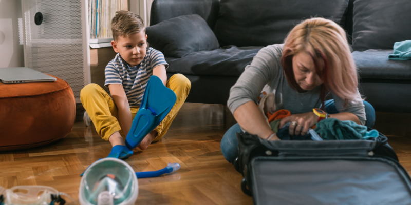 Mom packing son's stuff into his luggage with swimming gear around them