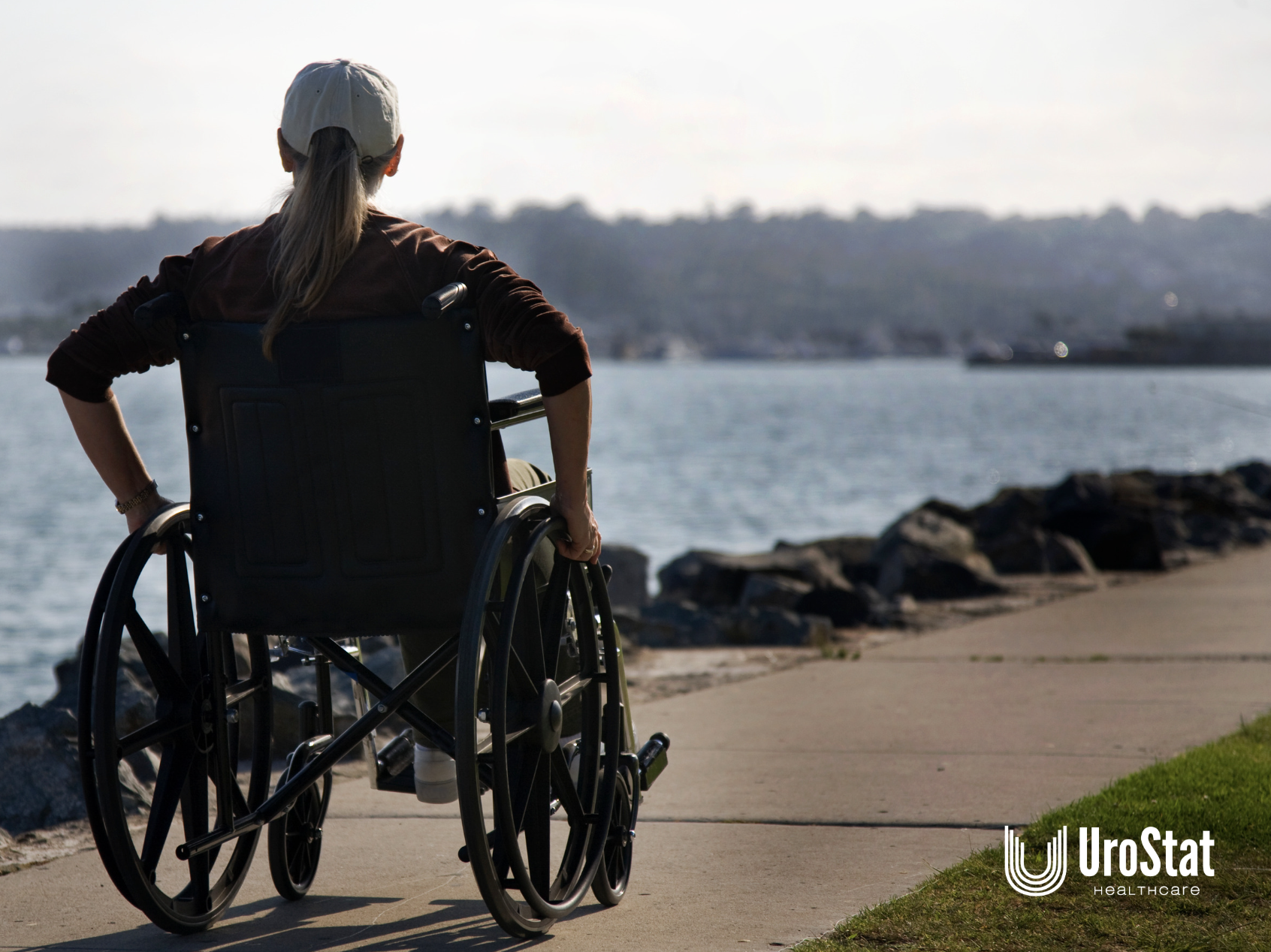 A woman using a wheelchair looks out over the water from a paved path.